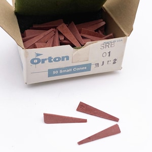 May include: A cardboard box labeled Orton 50 Small Cones containing numerous small, reddish-brown triangular ceramic cones. A green foam insert sits inside the open box, and three additional cones are placed on the surface in front.