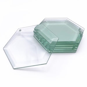 May include: A stack of six clear glass hexagon-shaped coasters. The coasters are slightly frosted and have a smooth, polished finish.