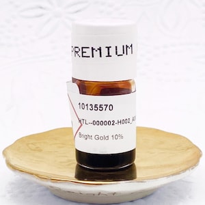 May include: A small glass bottle containing a dark liquid, topped with a white cap labeled PREMIUM. The bottle features a white label with the text 10135570, MTL--000002-H002_A, and Bright Gold 10%. It rests on a small, round, gold-rimmed ceramic dish.