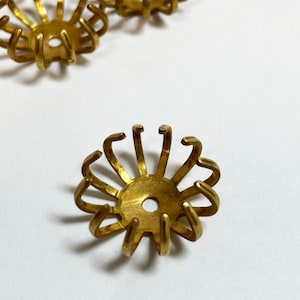 May include: A close-up of a group of gold-colored metal bead caps. The caps have a circular base with multiple curved prongs extending upwards. The prongs are arranged in a circular pattern, creating a flower-like shape.