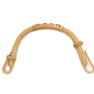 Può includere: Un manico curvo per borsa in rattan marrone chiaro con una sezione centrale intrecciata e dettagli annodati. Il manico ha anelli ad ogni estremità per il fissaggio. Lo sfondo è bianco.