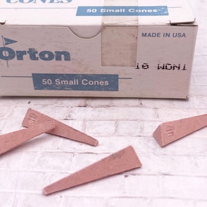 May include: A white cardboard box labeled "Orton Small Pyrometric Cones" with "50 Small Cones" printed on it. Several small, pink, triangular cones are scattered around the box. The box is marked "MADE IN USA."
