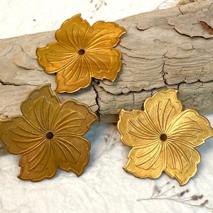 May include: Three gold metal flower-shaped embellishments with a raised center and intricate details. The embellishments are arranged on a piece of weathered wood.