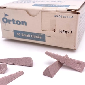 May include: A box of Orton brand 50 Small Cones, Made in USA. Several small, pinkish-purple cones are scattered in front of the box. The box is beige with blue text.