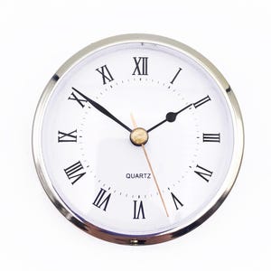 May include: A round quartz clock insert featuring a polished silver-tone metal bezel and a white face. The dial displays black Roman numerals for the hour markers, black hour and minute hands, and a thin gold-tone second hand. The word QUARTZ is printed.