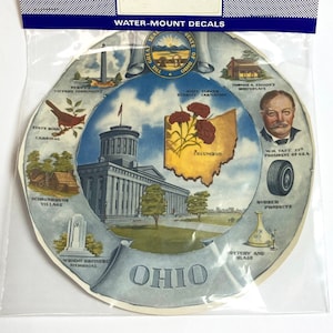 Könnte beinhalten: Ein Vintage-Wasser-Montage-Abziehbilder mit einer farbenfrohen Illustration des Bundesstaates Ohio. Das Abziehbilder enthält den Staatsvogel, den Kardinal, das State Capitol Building und die Staatsblume, die Nelke. Der Text "Ohio" ist prominent auf einem blauen Banner dargestellt.