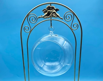 Puede incluir: Un soporte de metal dorado con una parte superior redonda y tres patas. Un globo de vidrio transparente cuelga de la parte superior del soporte.