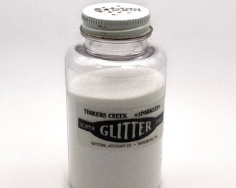 Puede incluir: Una botella de plástico transparente llena de purpurina blanca. La botella tiene una tapa de metal blanco con pequeños agujeros y una etiqueta que dice "Tinkers Creek Super Glitter Fine Sparkles". El producto es de National Artcraft Co., Twinsburg, OH.