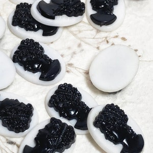 May include: A collection of oval-shaped cameo cabochons. Each cabochon features a black profile against a white background. The profile depicts a woman with detailed hair and facial features. These are likely for jewelry making or crafting.