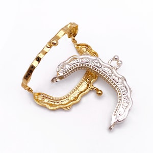 May include: Two ornate purse frames in gold and silver tones. The gold frame has a textured design, while the silver frame features intricate floral patterns. Both frames are curved and have a clasp mechanism, suggesting they are for a small bag or clutch.