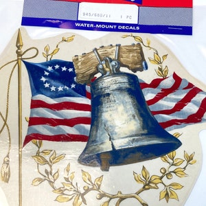 May include: A vintage water-mount decal featuring a Liberty Bell with a cracked surface, surrounded by a red, white, and blue American flag and gold-colored leaves. The decal is labeled "Color Brite" and "Water-Mount Decals".