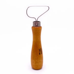 May include: A pottery loop tool with a wooden handle and a metal loop. The handle is light brown and has the text "KEMPER LTD USA" printed on it. The metal loop is silver and is used for shaping and trimming clay.
