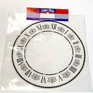 May include: A white clock face with black Roman numeral markings and a black rope design around the edge. The clock face is labeled "Color Brite" and "Water-Mount Decals".