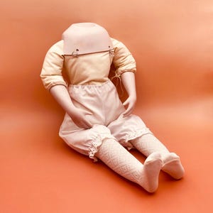 Peut inclure: Une poupée vintage avec un corps crème, vêtue d'un haut jaune clair, d'un bloomer blanc et de bas à motifs. La tête de la poupée est manquante, révélant une douille de cou. La poupée est posée sur un fond orange.