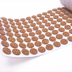 Puede incluir: Una hoja de c&iacute;rculos de corcho marr&oacute;n sobre un fondo blanco. Los c&iacute;rculos est&aacute;n dispuestos en filas y tienen un di&aacute;metro de aproximadamente 2,5 cm.