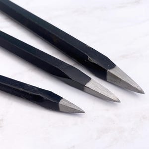Può includere: Tre scalpelli in metallo con lunghi e sottili manici esagonali rivestiti di nero e punte affilate in acciaio color argento. Gli utensili sono disposti in diagonale, mettendo in risalto il design affusolato e la struttura metallica.