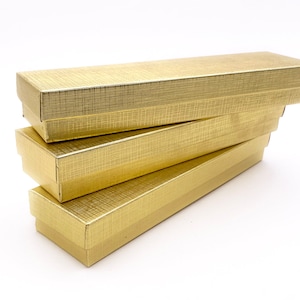 Pode incluir: Quatro caixas de cartão de cor dourada empilhadas umas sobre as outras. As caixas são retangulares e têm um padrão texturizado subtil.