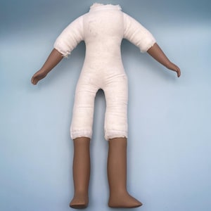 May include: A doll body with a white fabric torso, arms, and legs. The arms and legs are a dark brown colour. The doll's arms are outstretched, and the legs are straight. The doll has no head.