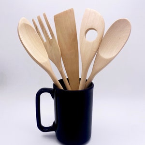 May include: Five wooden kitchen utensils, including a spoon, spatula, slotted spoon, and two forks, are standing upright in a dark blue ceramic mug.