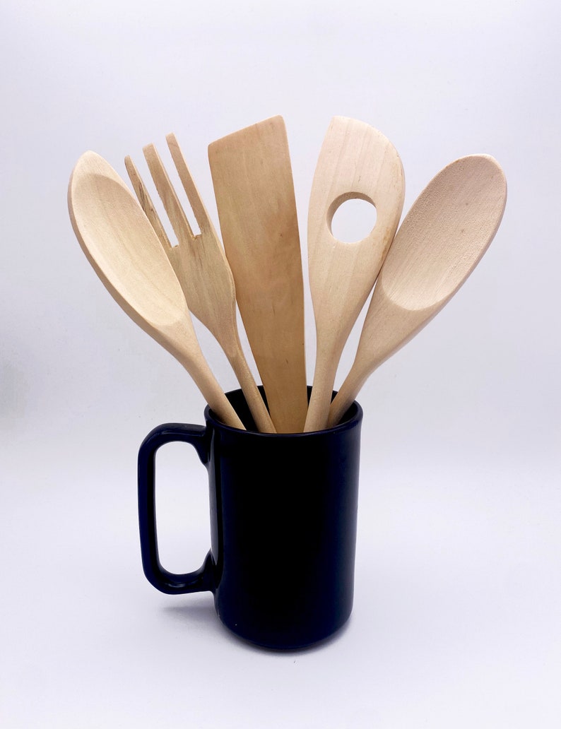 May include: Five wooden kitchen utensils, including a spoon, spatula, slotted spoon, and two forks, are standing upright in a dark blue ceramic mug.