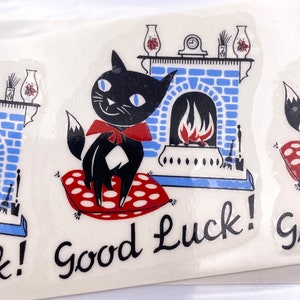 Może przedstawiać: Czarny kot z czerwoną kokardką siedzi na czerwono-białej poduszce w kropki przed kominkiem. Kominek ma niebieski ceglany wygląd zewnętrzny i palący się w nim ogień. Tekst "Good Luck!" jest napisany czarnymi literami pod kotem.