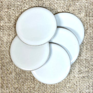 May include: Five white ceramic coasters, stacked in a circular pattern. The coasters are smooth and round, with a slightly raised edge.