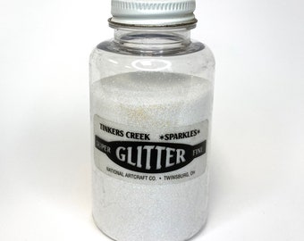 Puede incluir: Un frasco de plástico transparente lleno de purpurina blanca. La etiqueta dice "Tinkers Creek *Sparkles* Super Fine Glitter National Artcraft Co. Twinsburg, OH"