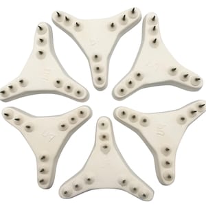 Pode incluir: Seis bases brancas em forma triangular com múltiplos pequenos pinos de metal em cada base. As bases estão dispostas em círculo e cada base tem as letras "BU" impressas.