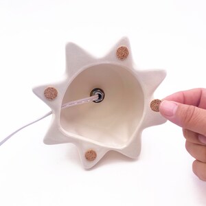 Puede incluir: Base de l&aacute;mpara de cer&aacute;mica blanca con un dise&ntilde;o de estrella y almohadillas de corcho en la parte inferior. La base de la l&aacute;mpara tiene un agujero en el centro para una bombilla.