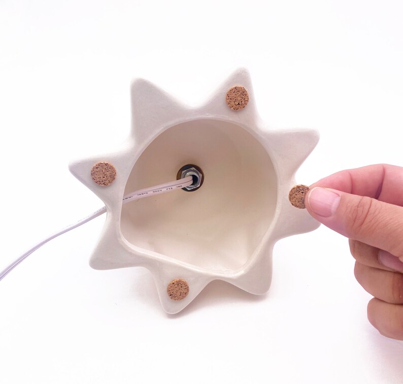Puede incluir: Base de l&aacute;mpara de cer&aacute;mica blanca con un dise&ntilde;o de estrella y almohadillas de corcho en la parte inferior. La base de la l&aacute;mpara tiene un agujero en el centro para una bombilla.