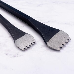 May include: Two metal tools with long, slender, black-coated hexagonal shafts and flat, silver-colored, multi-pronged heads. The heads feature a series of sharp, tooth-like projections designed for gripping or prying, with a distinct contrast between the dark handles and the metallic, textured tips.