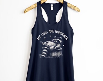May include: A navy blue tank top with a white graphic of a raccoon lying down with the text "My legs are hungover".