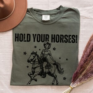 May include: Green t-shirt with a black graphic of a woman riding a horse. The graphic says "HOLD YOUR HORSES!"