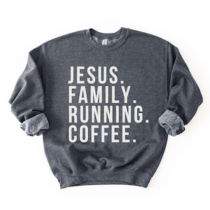 May include: A dark gray sweatshirt with the words "JESUS. FAMILY. RUNNING. COFFEE." printed in white, stacked vertically. The sweatshirt has a crew neck and long sleeves with rolled-up cuffs. The background is plain white.
