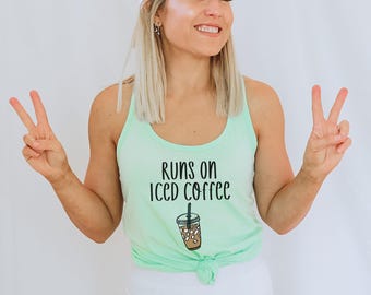 May include: A woman wearing a light green tank top with the text "runs on iced coffee" and a drawing of a cup of iced coffee. She is wearing a white baseball cap and white pants. She is smiling and giving the peace sign with both hands.