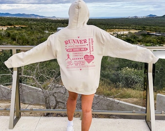 May include: A cream-colored hoodie with a pink graphic that reads "RUNNER" and "ONE MORE MILE." The design includes a running figure, a heart, and other text. The hoodie is worn with white shoes and a skirt.