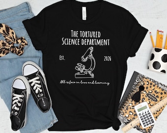 Pode incluir: Uma t-shirt preta com texto branco que diz The Tortured Science Department, Est. 2026, e All is fair in love and learning. Um desenho de linha branca de um microscópio está centrado entre os elementos de texto na frente da t-shirt.