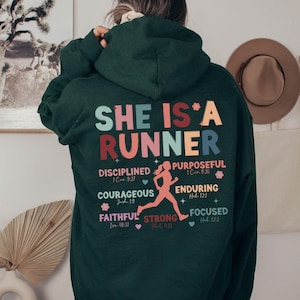 May include: A dark green hoodie with the text "SHE IS A RUNNER" in colorful letters. The hoodie also features a running woman graphic and inspirational quotes with bible verses.