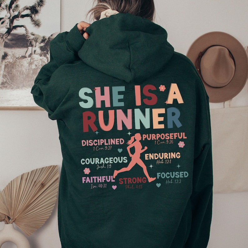 May include: A dark green hoodie with the text "SHE IS A RUNNER" in colorful letters. The hoodie also features a running woman graphic and inspirational quotes with bible verses.