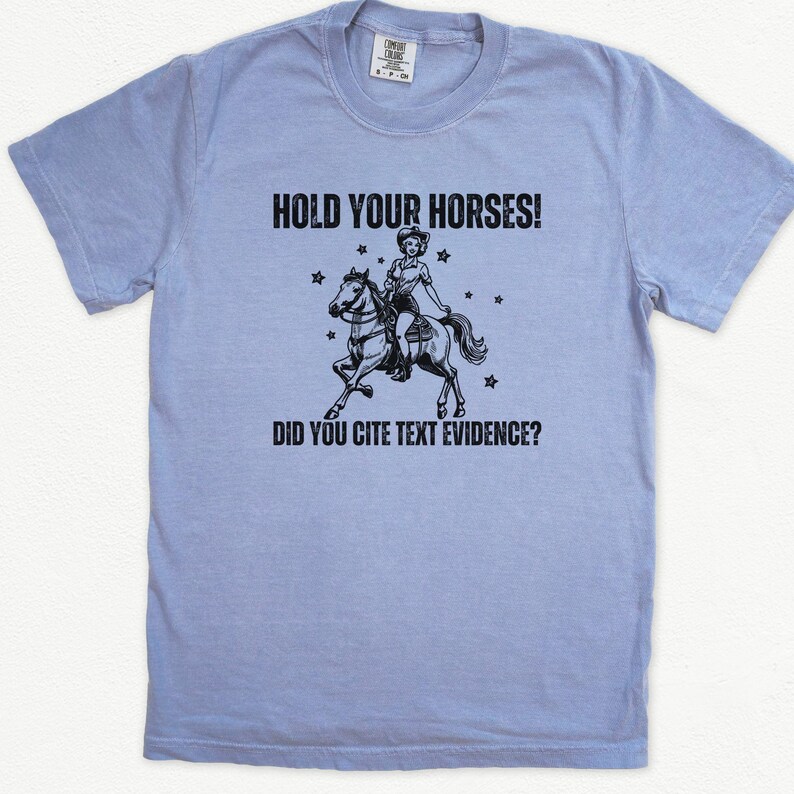 May include: A light blue t-shirt with a black graphic of a woman riding a horse. The text "Hold Your Horses! Did You Cite Text Evidence?" is printed on the shirt.