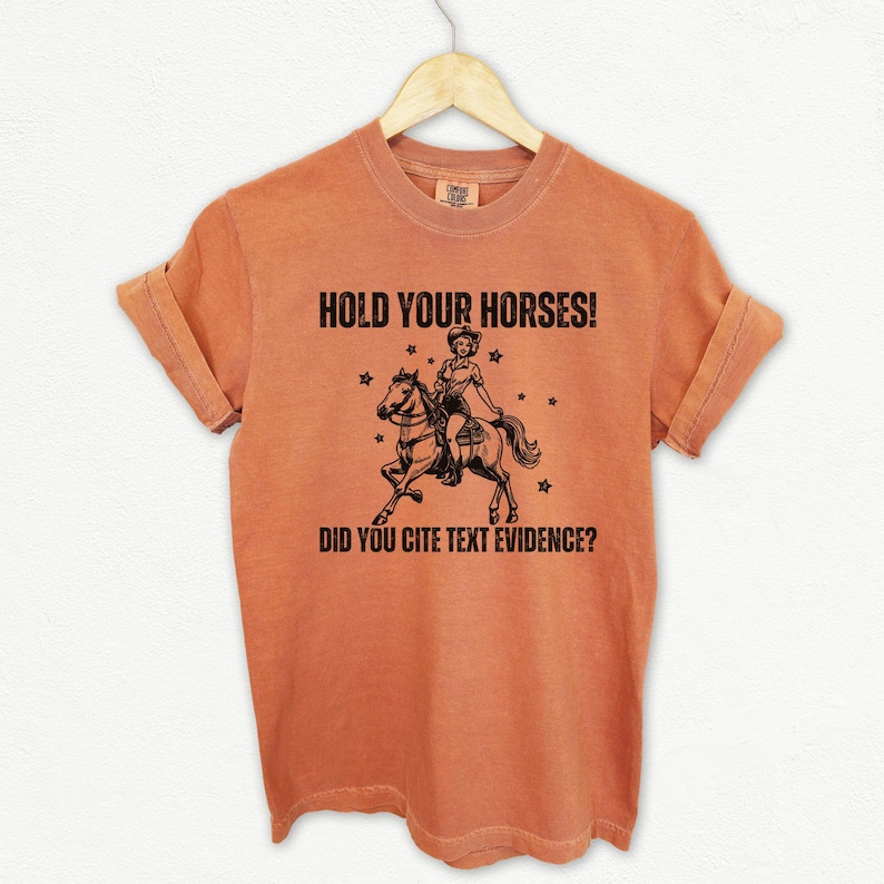 May include: A burnt orange t-shirt with the text "HOLD YOUR HORSES! DID YOU CITE TEXT EVIDENCE?" and a black and white illustration of a woman riding a horse.