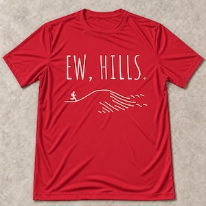 May include: A bright red short-sleeved t-shirt featuring white text that reads "EW, HILLS." and a white line drawing of a stick figure running up a steep hill. The design is centered on the chest of the shirt, which has a crew neckline.