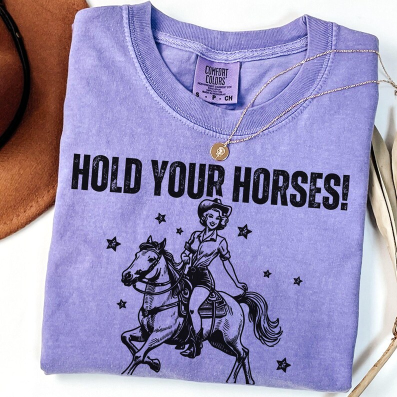 May include: Purple t-shirt with a black graphic of a woman riding a horse. The text "HOLD YOUR HORSES!" is printed on the shirt.