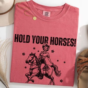 May include: A pink t-shirt with the text "HOLD YOUR HORSES!" in black. A black and white illustration of a woman riding a horse with stars in the background.