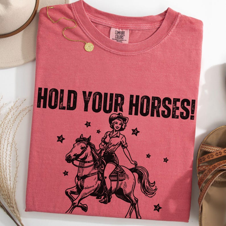 May include: A pink t-shirt with the text "HOLD YOUR HORSES!" in black. A black and white illustration of a woman riding a horse with stars in the background.