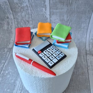 May include: A white cake decorated with a calculator, a red pen, and four stacks of colourful books. The books are red, orange, green, and blue.