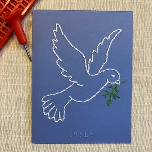 May include: A blue card with a white embroidered dove carrying a green olive branch. The card has braille text at the bottom.
