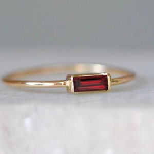 Könnte beinhalten: Ein zarter Goldring mit einem rechteckigen, tiefroten Edelstein. Der Edelstein ist horizontal in einer goldenen Lünette gefasst, was ein minimalistisches und elegantes Design ergibt. Der Ring hat ein dünnes, poliertes Band, das den Fokus auf den leuchtend roten Stein lenkt.