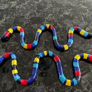 May include: Two colourful, serpentine glass sculptures. Each snake-like form is segmented with alternating bands of blue, yellow, red, and black. The sculptures are displayed on a dark, speckled surface.