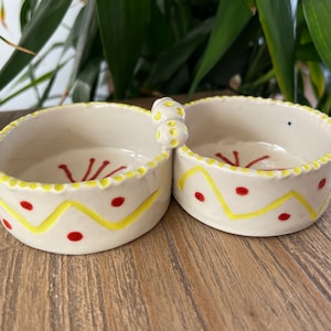 May include: A white ceramic double dish with a yellow and red geometric design. The two dishes are connected by a small handle.